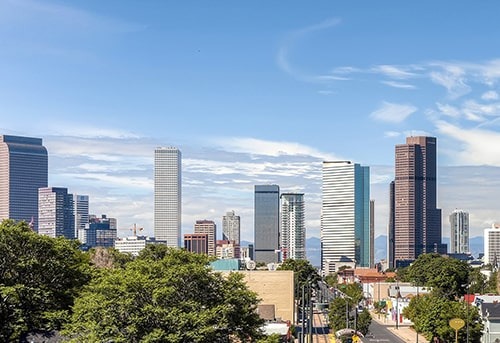 Vibrant skyline of Denver, Colorado with modern skyscrapers and trees in the foreground against a blue sky.