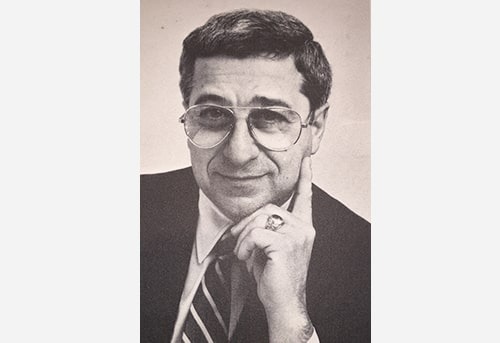 A middle-aged man with dark hair and glasses, wearing a suit and tie, looking directly at the camera.