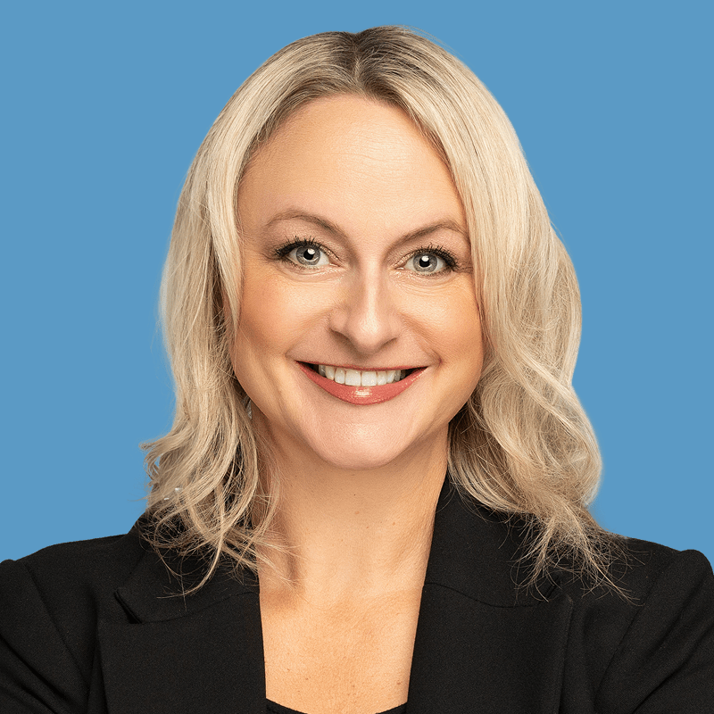 A smiling woman with blonde hair wearing a black jacket against a blue background.