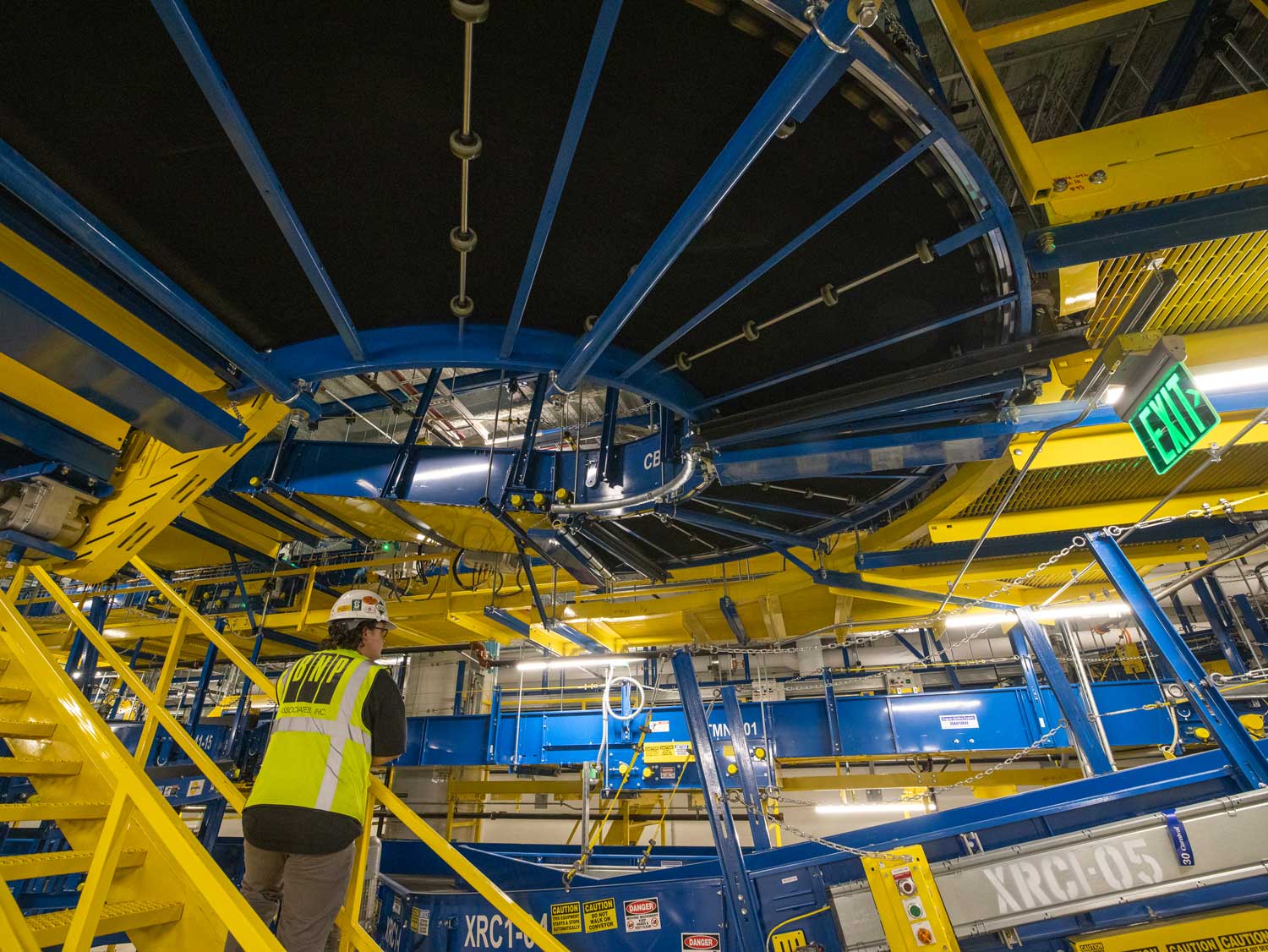 Vast industrial interior with complex network of blue and yellow metal beams, machinery, and an employee in protective equipment.