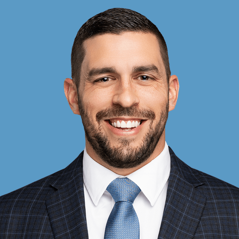 Headshot of a smiling man with a beard, wearing a navy blue checkered suit and blue tie, set against a bright blue background.