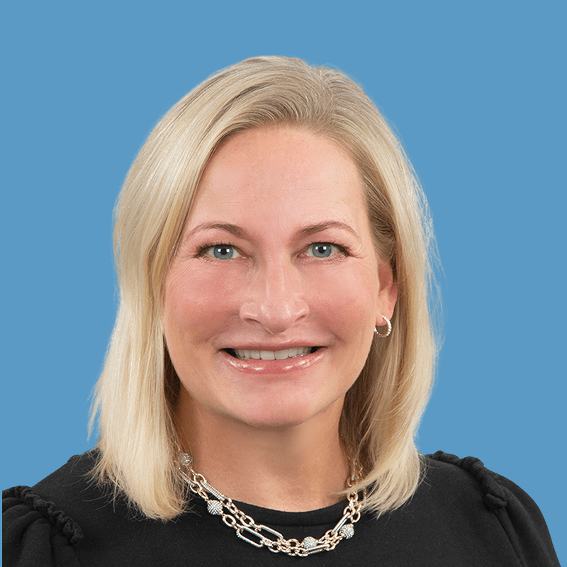 Smiling woman with blonde hair and a black top wearing a chunky necklace against a blue background.