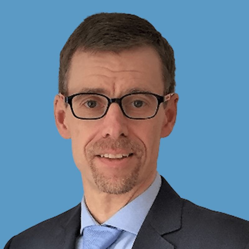 Headshot of a middle-aged man with short brown hair, glasses, and a blue tie, smiling against a blue background.