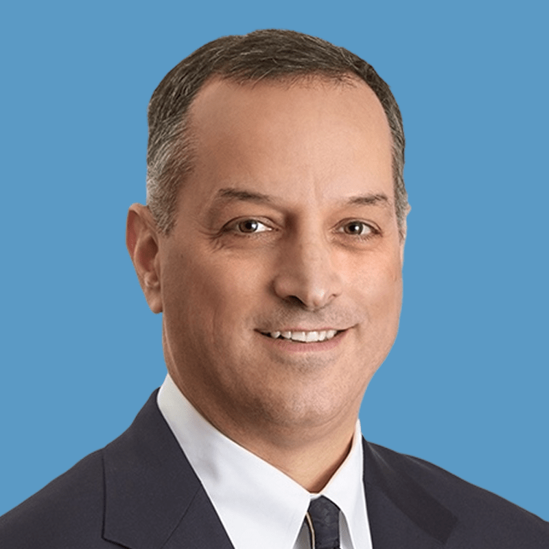 Headshot of a smiling middle-aged man in a suit against a blue background.