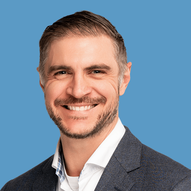 Smiling man with gray hair and beard wearing a suit against a blue background.
