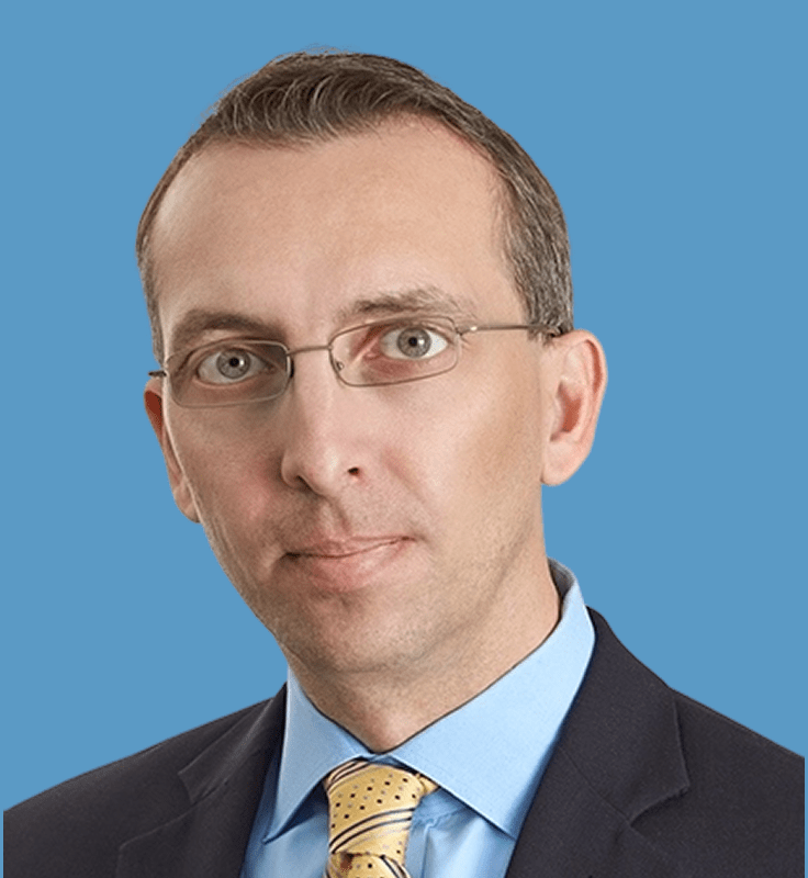 Portrait of a man wearing glasses, a suit, and a patterned tie against a blue background.