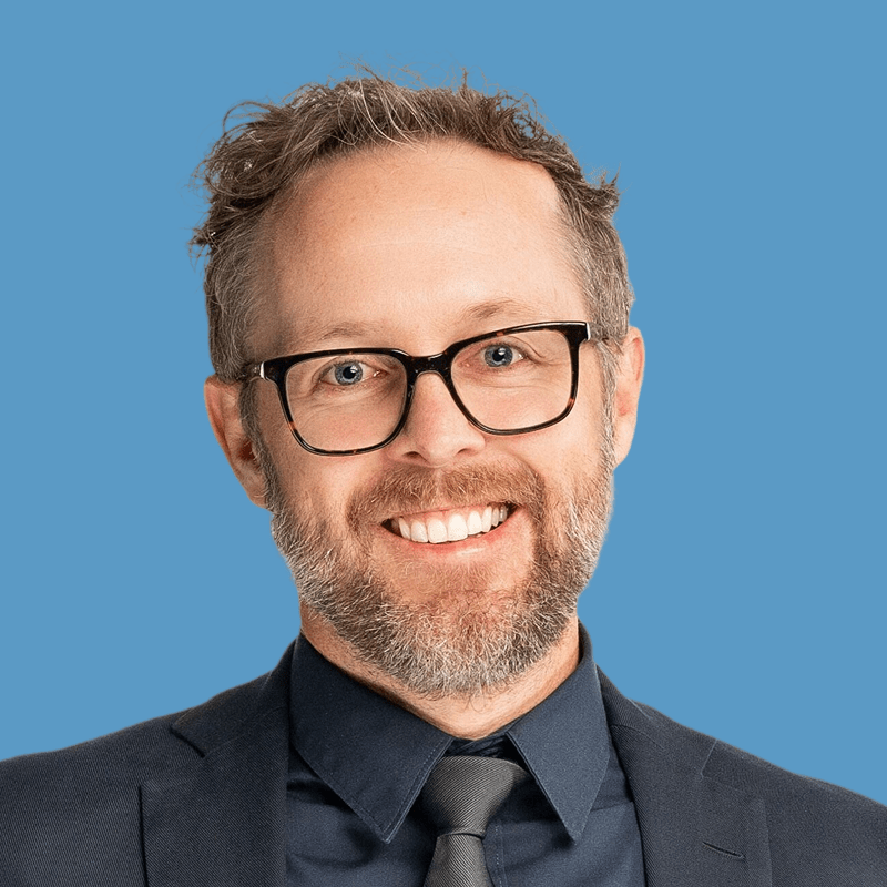 Smiling man with beard and glasses wearing a suit and tie against a blue background.